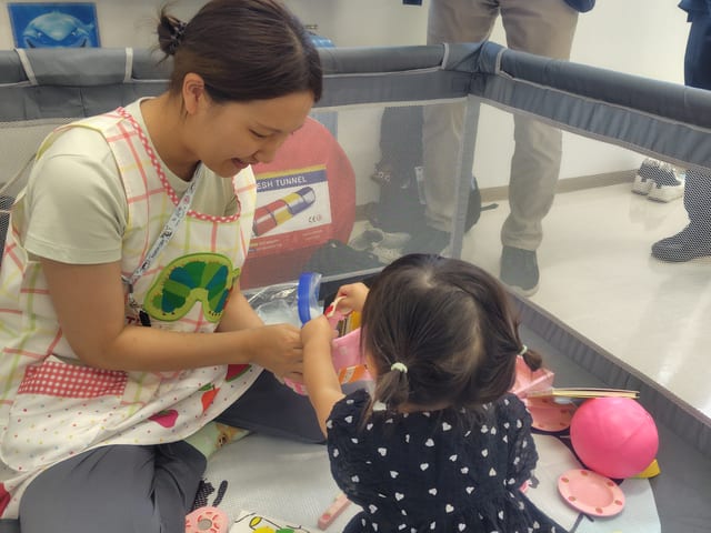mama's marche(相浦コミュニティセンター)の託児ブースで、スタッフが笑顔で子どもと一緒におもちゃで遊んでいる様子